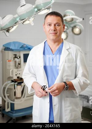 Portrait eines Mannes Chirurgen, der die Kamera anschaut, im Operationssaal im Krankenhaus stehend. Schöner männlicher Arzt trägt weißen Laborkittel. Konzept der Medizin, medizinisches Personal und plastische Chirurgie.