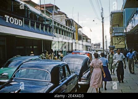 Ein Blick auf Fußgänger und alte Autos in Frederick Street, Port of Spain, Trinidad und Tobago c 1958. Die Straße verläuft nach Norden durch die Stadt im zentralen Geschäftsviertel der Innenstadt. Das Downtown Viertel von Port of Spain ist auch der älteste Bezirk der Stadt. Frederick Street bot traditionelle Kolonialarchitektur und einige berühmte alte Geschäfte, wie die Miller's, Todd und Hendinning Geschäfte hier. Dieses Bild ist von einem alten Amateur-35mm-Farbtransparenz. Stockfoto