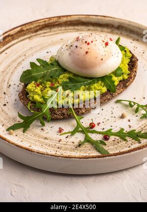 Vertikale Nahaufnahme pochiertes Eiertoast mit Avocado, gesalzenem Lachs, Rucola und Roggenbrot auf Keramikplatte, gesundes Lebensmittelkonzept Stockfoto