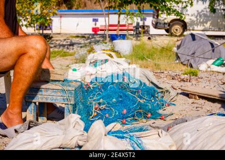 Ein Fischer sitzt auf einem Stuhl am Strand und reinigt Fischernetz von Muscheln, zerschlagen sie mit einem Holzbrett. Stockfoto