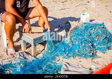 Ein Fischer sitzt auf einem Stuhl am Strand und reinigt Fischernetz von Muscheln, zerschlagen sie mit einem Holzbrett. Stockfoto