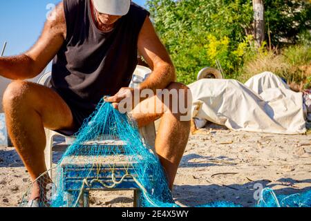 Ein Fischer sitzt auf einem Stuhl am Strand, stapelt Fischernetz auf und reinigt es von Muscheln. Stockfoto