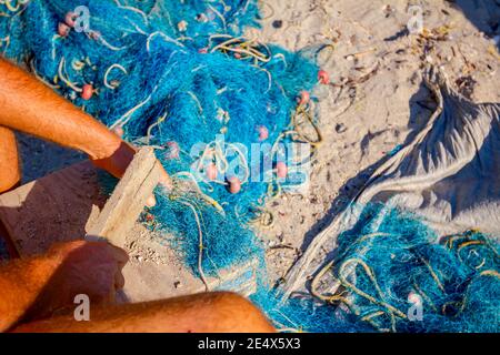 Ein Fischer sitzt auf einem Stuhl am Strand und reinigt Fischernetz von Muscheln, zerschlagen sie mit einem Holzbrett. Stockfoto