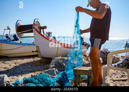 Fischer häufen Fischnetz und bereiten sich auf sein nächstes Angeln. Die Boote sind trocken am Sandstrand angedockt. Stockfoto