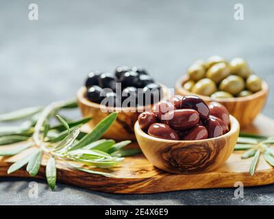 Set aus grünen, roten und schwarzen Oliven auf grauem Hintergrund. Verschiedene Olivenarten in Olivenholzschüsseln und Olivenöl über Holzschneidebrett und frischen Olivenblättern. Speicherplatz kopieren. Stockfoto