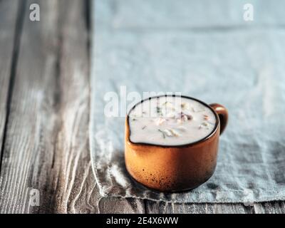 Authentische pakistanische Gurken-Raita-Sauce auf blauer Leinenserviette auf grauem Holzhintergrund. Raita Dip mit Gurken, Zwiebeln und Tomaten Add-ins in braunem Keramik-Sauceboat. Speicherplatz kopieren Stockfoto