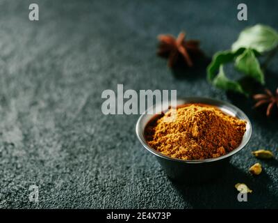 Indische oder pakistanische Masala Pulver und Gewürze auf dunklen strukturierten Hintergrund. Kleine Metallschüssel mit trockenem Curry Garam Masala Mix Gewürzmischung, grünem Lorbeerblatt, Kardamom und Sternanis mit Kopieplatz links Stockfoto