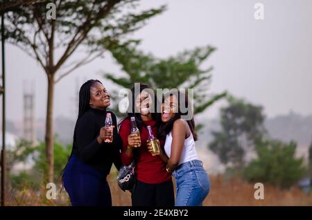 Junge schöne afrikanische Damen machen einen Spaziergang in einem Garten, halten Flaschen von Getränken und mit glücklichen Diskussion Stockfoto