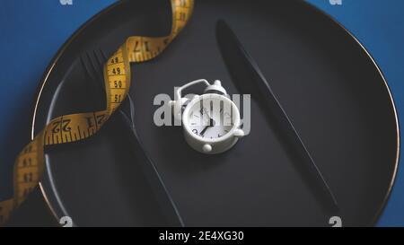 Nahaufnahme von Gabel und Küchenmesser auf einer Platte mit Maßband und weißem Wecker. Intermittierende Fasten Konzept Diät gesunde Lebensweise Stockfoto