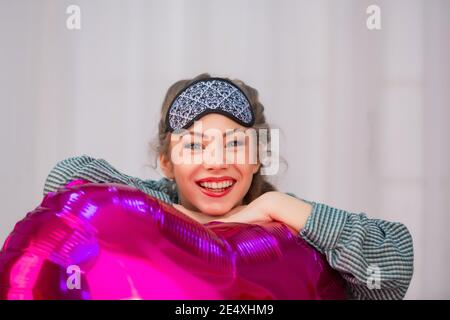 Ein junges gesundes Mädchen in einer Schlafmaske lächelt und umarmt einen rosa Ballon in Form eines Herzens, eines Portraits, eines Ortes für Text. Hochwertige Fotos Stockfoto