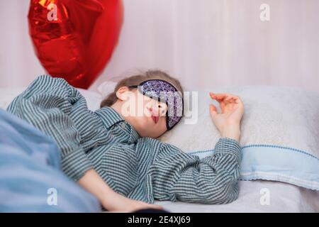 Eine junge gesunde Frau schläft im Bett, trägt eine Schlafmaske und Schlafanzug, im Hintergrund ein Ball in der Form eines roten Herzens. Hochwertige Fotos Stockfoto