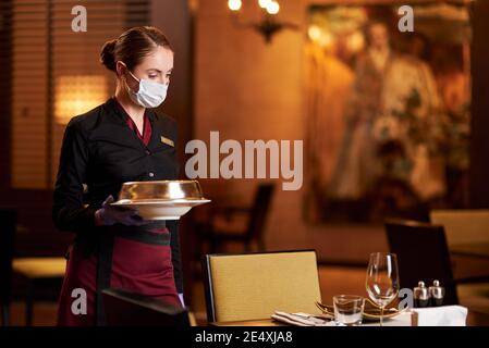 Kopieren Raum Foto der achtsamen Kellnerin trägt Schutzmaske während Essen auf den Tisch bringen Stockfoto