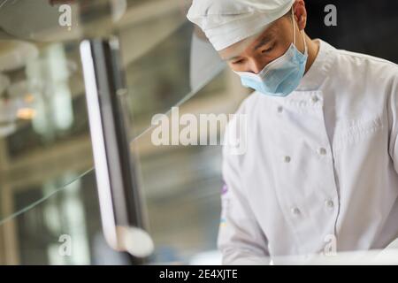 Zugeschnittenes Foto von jungen asiatischen Mann in Koch Uniform tragen Gesichtsmaske und Blick nach unten Stockfoto