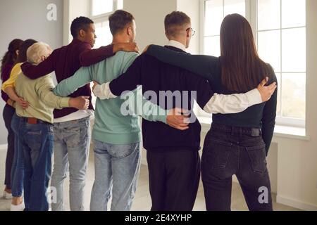 Eine Gruppe verschiedener Menschen steht in einer Umarmung mit dem Rücken zur Kamera und schaut aus dem Fenster. Stockfoto