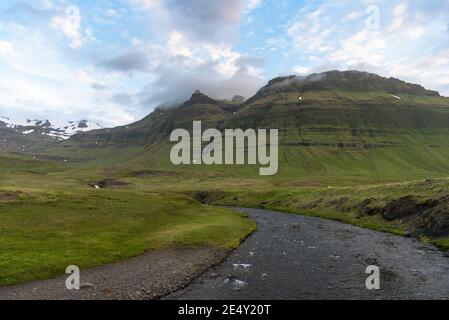 Fluss, der an einem Sommerabend durch ein grünes Tal fließt, umgeben von hoch aufragenden, grasbewachsenen Bergen. Schneebedeckte Gipfel sind in der Ferne sichtbar. Stockfoto