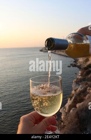 Nahaufnahme eines Mannes, der Weißwein in einen eingießt glas mit einer wunderschönen Landschaft der Bozcaada Insel bei Sonnenuntergang Im Hintergrund Stockfoto