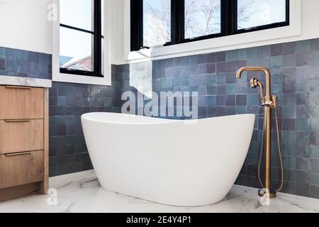 Ein wunderschönes, luxuriöses Detail im Badezimmer, das von einer weißen, eigenständigen Badewanne und einem goldenen Wasserhahn auf weißen Marmorfliesen und vor einer blau gefliesten Wand aufgenommen wurde. Stockfoto