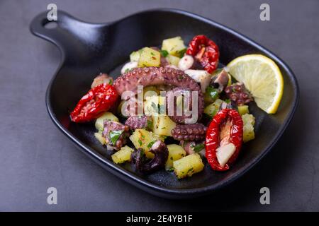 Warmer Salat mit Oktopus, Kartoffeln, getrockneten Tomaten, Knoblauch und Zitrone in einer schwarzen kleinen Pfanne. Nahaufnahme, schwarzer Hintergrund. Stockfoto