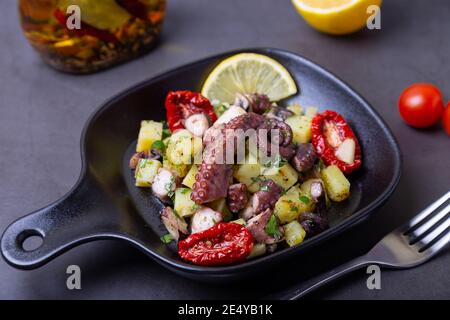 Warmer Salat mit Oktopus, Kartoffeln, getrockneten Tomaten, Knoblauch und Zitrone in einer schwarzen kleinen Pfanne. Nahaufnahme, schwarzer Hintergrund. Stockfoto