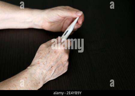 Ältere Frau misst Körpertemperatur, Konzept von Fieber, Coronavirus-Symptome. Digitalthermometer in faltigen weiblichen Händen aus der Nähe Stockfoto