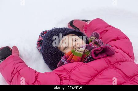 Ein kleines Mädchen in einer rosa Jacke macht einen Engel auf dem frisch gefallenen Schnee. Winter Kinder Spaß Konzept. Stockfoto