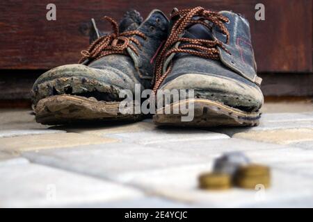 Abgenutzte Stiefel mit einer nachlagenden Sohle auf einem Hintergrund aus grauen Fertigern und dunkelbraunem Holz. Im Vordergrund ist ein verschwommener Stapel von Münzen. Das Konzept des Povers Stockfoto