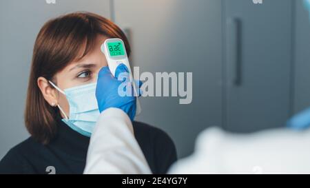 Kaukasischer Arzt trägt Handschuhe misst die Körpertemperatur von Der Patient mit Maske während einer Konsultation Stockfoto