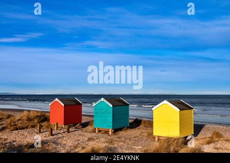 FINDHORN MORAY KÜSTE SCHOTTLAND DER STRAND MIT DREI FARBIGEN CHALETS ODER HÜTTEN IM WINTER UND SCHNEE AUF DEN HÜGELN Stockfoto