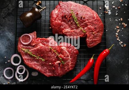 Zwei Rohe Rindersteaks mit Gewürzen, Zwiebeln und Chili auf dunklem Schiefer oder Beton. Draufsicht Stockfoto