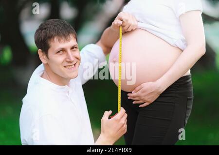 Ein junger Mann lächelt und misst den Magen seiner schwangeren Frau Mit einem Maßband in einem Sommerpark Stockfoto