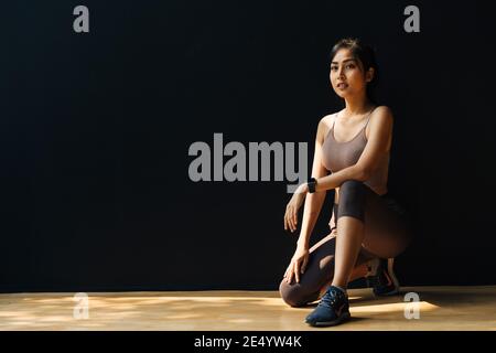 Fokussierte junge asiatische Sportlerin entspannen und sich auf das Training vorbereiten Beim hocken im Studio auf schwarzem Hintergrund - Ganzkörper Länge Stockfoto