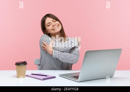 Egoistische glückliche Frau Büroangestellte umarmt sich mit Vergnügen Ausdruck, sitzen am Arbeitsplatz, Gefühl stolz und zufrieden. Innenaufnahme des Studios ist Stockfoto