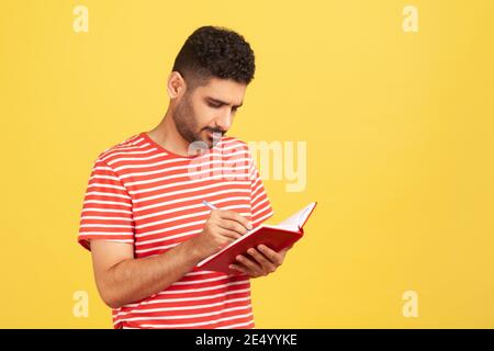 Ernst konzentrierter Geschäftsmann mit Bart in gestreiftem T-Shirt notieren Gedanken halten Tagebuch, Notizen auf Notizblock. Innenaufnahme des Studios isola Stockfoto