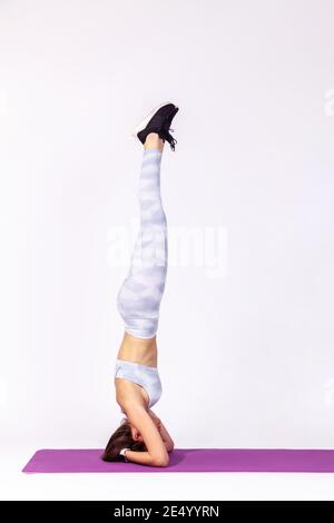 In voller Länge Seitenansicht athletische Frau in weißer Sportbekleidung dabei Yoga Kopfstand Pose auf Matte, Gymnastik für Körper Gleichgewicht und Flexibilität. Studio im Innenbereich Stockfoto