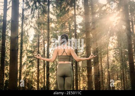 Frau, die bei Sonnenaufgang in einem Wald steht, Yoga praktiziert und Sportkleidung meditiert. Stockfoto