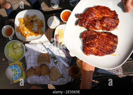 Zwei frisch gegrillte Schweinelende-Steaks auf weißem Teller, von Hand über den Gartentisch gehalten, mit mehr Essen vom Grill. Stockfoto