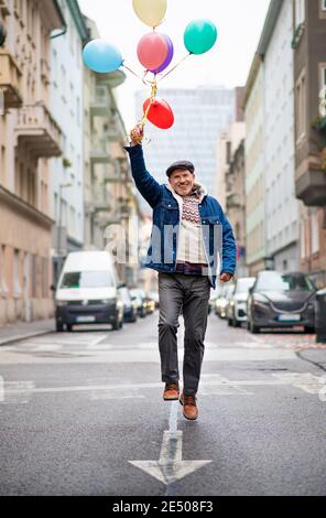 Glücklicher älterer Mann mit Luftballons draußen auf der Straße in der Stadt, zu Fuß. Stockfoto