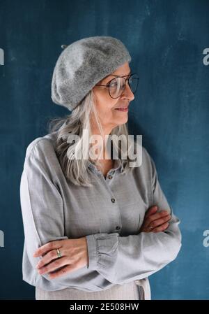 Porträt einer älteren Frau mit grauer Baskenmütze, die vor dunklem Hintergrund im Innenbereich steht. Stockfoto