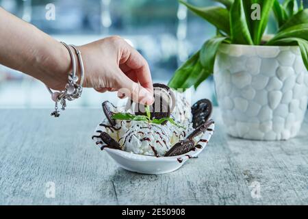 Eine Frau Hand hält ein Stück Oreo mit Eis, Schokoladensirup auf der weißen Platte im Marmorhintergrund Stockfoto