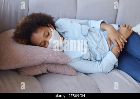 Gemischtes Rennen Teenager-Mädchen in legerer Kleidung tragen schützende Gesichtsmaske entspannend, liegend auf der Couch, während die Zeit zu Hause während der Quarantäne Stockfoto