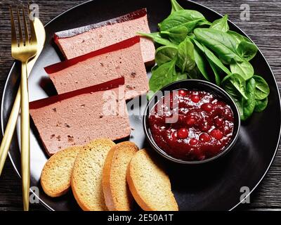 Französisches Gericht Entenleber Pastete mit Cranberry Marmelade auf einem schwarzen Teller serviert mit Cranberry-Sauce, Spinat und Brioche Croutons, mit goldenem c Stockfoto