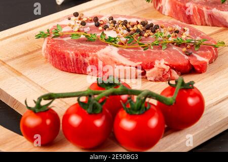 Frisch gehackte rohe Schweinesteaks mit Gewürzen, reifen Tomaten und Thymian auf einem Schneidebrett auf einem schwarzen Holztisch Stockfoto