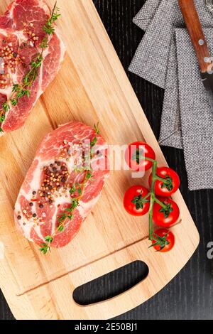 Frisch gehackte rohe Schweinesteaks mit Gewürzen, Tomaten und Thymian auf einem Schneidebrett auf einem schwarzen Holztisch. Neben einer Serviette mit Messer und Fo Stockfoto