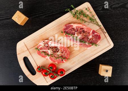 Frisch gehackte rohe Schweinesteaks mit Gewürzen, Tomaten und Thymian auf einem Schneidebrett auf einem schwarzen Holztisch. Neben Pfefferstreuer und Salz schütteln Stockfoto