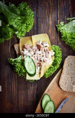 Flachlage Nahaufnahme von Sandwich mit grünen Salatblättern, Käse, Thunfisch, Gurken und Weizenbrot, auf Holz dunkel rustikalen Tisch Hintergrund, Stockfoto