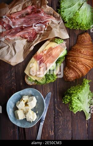 Rustikale Küche Holztisch mit Sandwich Croissant, Marmelade Fleisch Scheiben, Hartkäse, grünen Salatblättern, frische Gurken, Butter und Messer, Flatlay, Stockfoto
