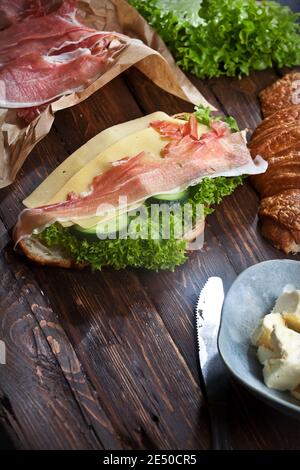 Croissant-Sandwich mit Marmelade-Fleischscheiben, grünen Salatblättern, frischen Gurken, Käse, Butter und Messer, auf rustikalem Holztisch, flachgeebert Stockfoto