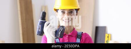 Frau Baumeister in Overalls und Schutzhelm halten Drill in Hands in Workshop Stockfoto