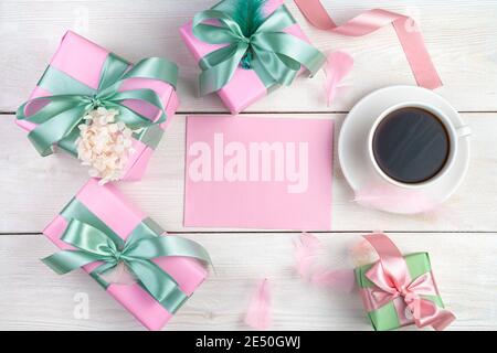 Eine festliche Komposition mit Geschenken, Kaffee und einem Umschlag in rosa Tönen auf hellem Holzhintergrund. Stockfoto
