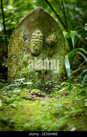 Nahaufnahme eines niedrigen, alten und schrägen japanischen Grabsteins, bedeckt mit Moos, der zwischen grüner Vegetation liegt und mit dem Basrelief eines buddha graviert ist Stockfoto